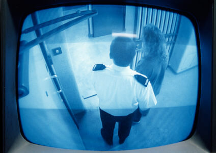 A TV monitor shows a police officer leading a detainee to a police cell.