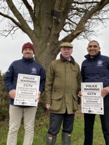 Rural Crime Advisors Bob Church (left) and Baz Bains (right) with Warwickshire Police and Crime Commissioner Philip Seccombe at Umberslade Road, near Earlswood, where one of the CCTV cameras is sited. A camera is fitted to the tree behind while the Rural Crime Advisors hold up signs warning of the CCTV coverage.