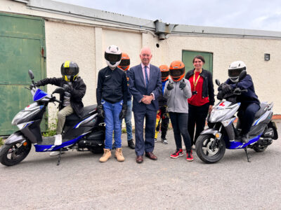 The Commissioner stands with a group of young moped riders in helmets.